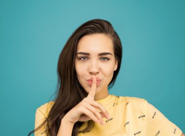 Portrait Photo of Woman in Yellow T-shirt Doing the Shh Sign While Standing In Front of Blue Background